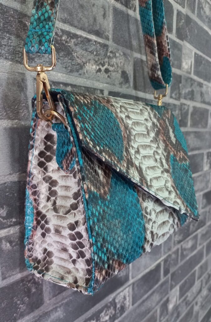 A snake skin crossbody purse in real Python leather with grey and turquoise colour. The bag hangs from a wall made of grey bricks.