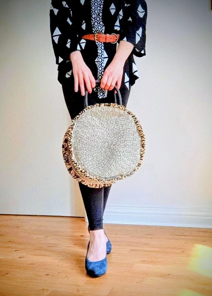 A woman standing indoors with a tote bag made of straw and with blue suede pumps. Surrounded by a white wall and wooden floor